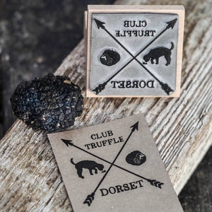 May include: A brown wooden stamp with black ink reads "Club Truffle Dorset" with a black and white illustration of a dog sniffing a truffle. The stamp is on a wooden surface next to a black truffle.