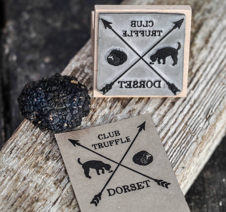 May include: A brown wooden stamp with black ink reads "Club Truffle Dorset" with a black and white illustration of a dog sniffing a truffle. The stamp is on a wooden surface next to a black truffle.