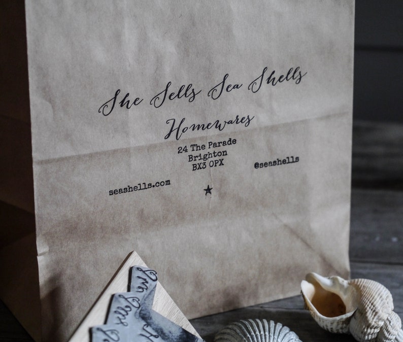 K&ouml;nnte beinhalten: Ein brauner Papierbeutel mit einem Logo, das "She Sells Sea Shells Homewares" und die Adresse "24 The Parade Brighton, BX3 OPX" sowie die Website "seashells.com" lautet. Das Logo ist in schwarzer Kursivschrift. Die Adresse und die Website sind in schwarzer serifenloser Schrift.