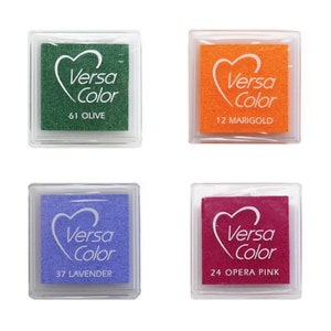 May include: Four ink pads in clear plastic cases. The ink pads are in the colours green, orange, blue, and pink. The ink pads are labelled with the brand name "Versa Color" and the colour name. The ink pads are 61 Olive, 12 Marigold, 37 Lavender, and 24 Opera Pink.