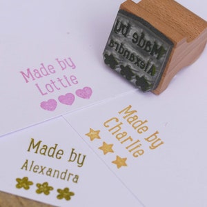 以下が含まれることがあります： 異なるデザインの3つの木製スタンプ。最初のスタンプは「Made by Lottie」と3つのピンクのハート。2番目のスタンプは「Made by Charlie」と3つの金の星。3番目のスタンプは「Made by Alexandra」と3つの金のフラワーです。