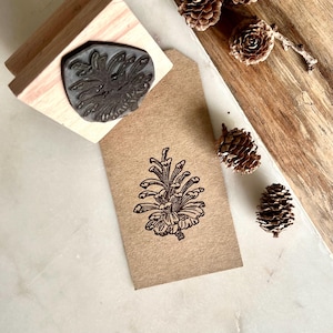 May include: A wooden stamp featuring a pine cone design, alongside a stamped kraft paper tag. The stamp is made of wood and rubber. Several pine cones are scattered around the tag on a wooden surface.