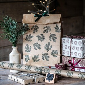 May include: A collection of holiday gift wrapping supplies. A brown paper bag and wrapping paper are printed with green leaf patterns. Wrapped gifts are stacked, and a sprig of pine is in a vase. A wooden stamp is also present.