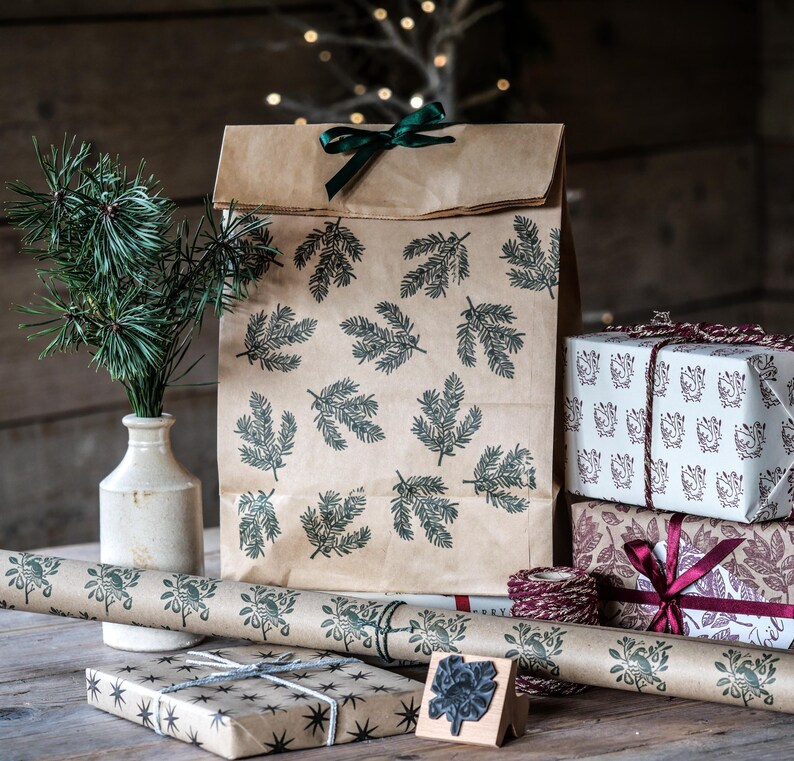 May include: A collection of holiday gift wrapping supplies. A brown paper bag and wrapping paper are printed with green leaf patterns. Wrapped gifts are stacked, and a sprig of pine is in a vase. A wooden stamp is also present.