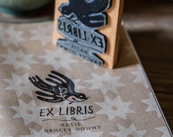 Può includere: Un foglio di carta marrone con stelle bianche stampate su di esso. Un timbro di gomma nero e bianco con un uccello e il testo "Ex Libris Katie Dancey-Downs" è sopra la carta.