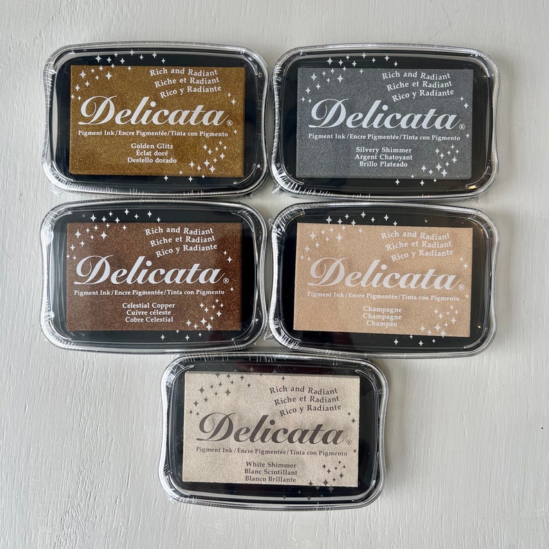 May include: Five black ink pad containers with gold, silver, copper, champagne, and white shimmer pigment ink. Each container has a label with the text "Delicata" and "Rich and Radiant" in English, French, and Spanish.