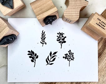 May include: Wooden stamps with botanical designs, including leaves and branches, are arranged around a white paper with matching black ink impressions. The stamps have wooden handles and the text "THE ENGLISH STAMP COMPANY" is visible on one stamp.