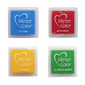 Könnte beinhalten: Vier Stempelkissen in durchsichtigen Plastikbehältern. Die Stempelkissen sind in den Farben Cyan, Scharlachrot, Kanariengelb und Frischgrün. Jedes Stempelkissen trägt den Aufdruck "Versa Color" sowie eine Nummer und den Farbnamen.