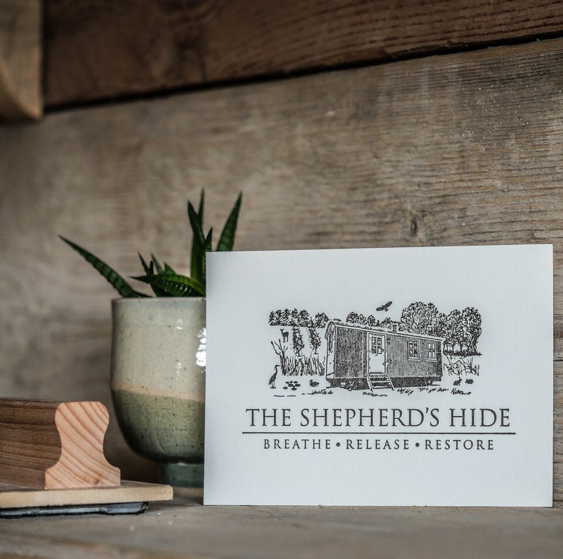 May include: A white card with a black and white illustration of a small cabin in a field. The text "The Shepherd's Hide" is printed below the illustration, with the tagline "Breathe. Release. Restore" printed below that.