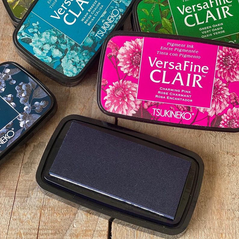 May include: A close-up of a black ink pad for stamping. The ink pad is labeled "VersaFine Clair" and "Charming Pink" in white text. The ink pad is made by Tsukineko.