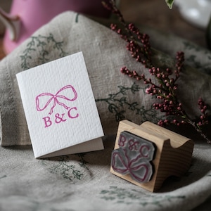Op de afbeelding: Een witte gevouwen kaart met een roze gestempelde afbeelding van een strik boven de letters B&C, vergezeld door een houten rubberen stempel met hetzelfde ontwerp. De stempel heeft een lichthouten handvat en een doorzichtige rubberen basis.