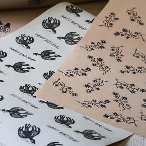 Puede incluir: Dos hojas de papel de regalo decorativo. Una es blanquecina con un patrón repetido de pájaros negros y el texto "HAPPY BIRTHDAY". La otra es de color canela con un diseño floral repetido en negro. El papel es probablemente para regalos.