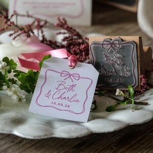 Op de afbeelding: Een wit rechthoekig papieren label met een roze lint, voorzien van een roze gedrukte rand met een strik en de tekst "Beth & Charlie 16.09.28". Daarnaast ligt een houten rubberstempel met hetzelfde ontwerp, inclusief de namen en datum.