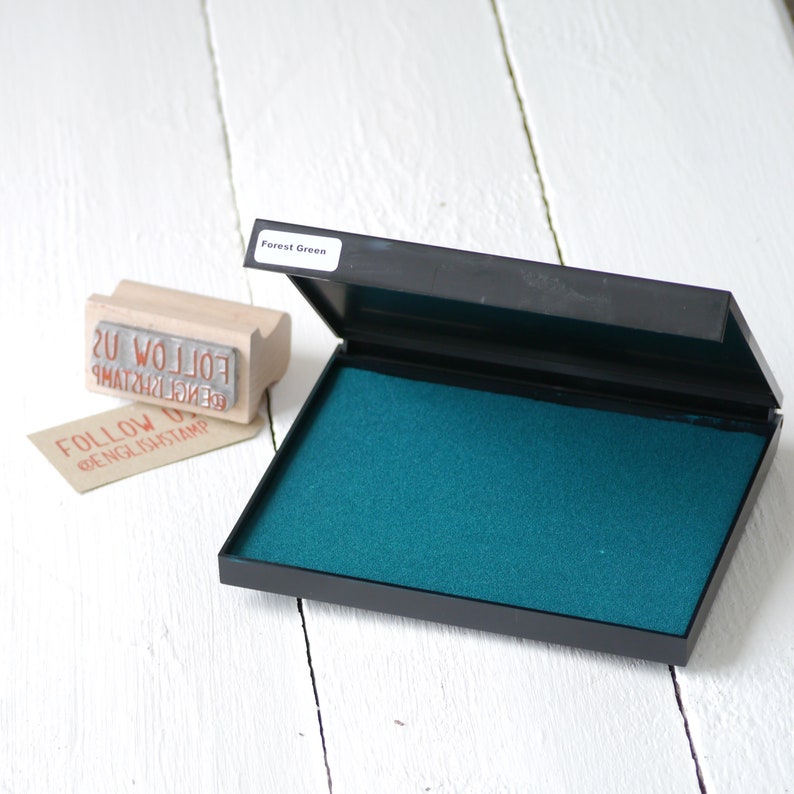 May include: An open ink pad with a dark green ink pad and a label that says "Forest Green". The ink pad is on a white wooden surface. There is a wooden stamp with the words "FOLLOW US" on it.