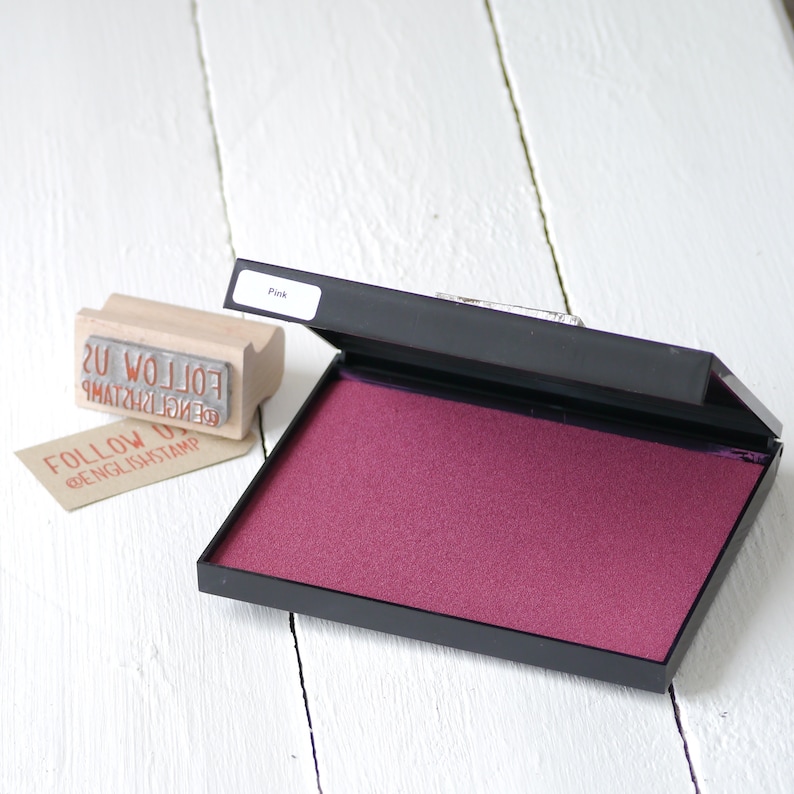 May include: An open ink pad with a pink ink pad inside. The ink pad is labeled "Pink". The ink pad is on a white wooden surface. The ink pad is used for stamping.