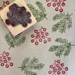 Op de afbeelding: Een houten stempel met een bessenontwerp wordt getoond naast een patroon van gestempelde bessen en dennentakken. De bessen zijn dieprood en de takken groen. De achtergrond is lichtbruin.