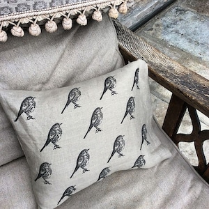 May include: A beige linen throw pillow with a black and white repeating pattern of birds. The pillow is sitting on a beige linen couch.