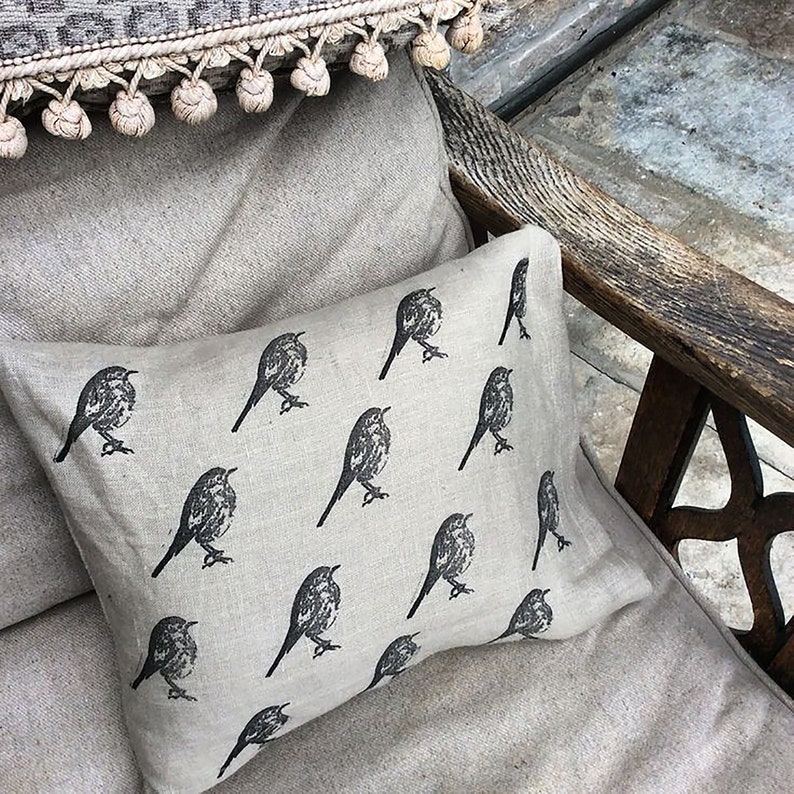 May include: A beige linen throw pillow with a black and white repeating pattern of birds. The pillow is sitting on a beige linen couch.