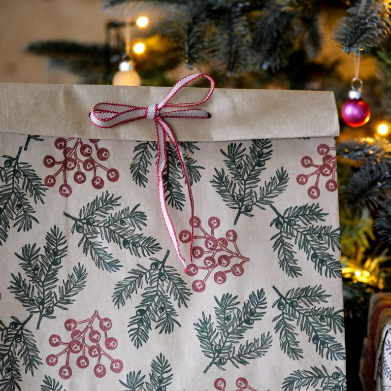 May include: A brown paper gift bag with a festive pattern of green pine branches and red berries. The bag is tied with a white and red ribbon. The background includes a Christmas tree with ornaments.