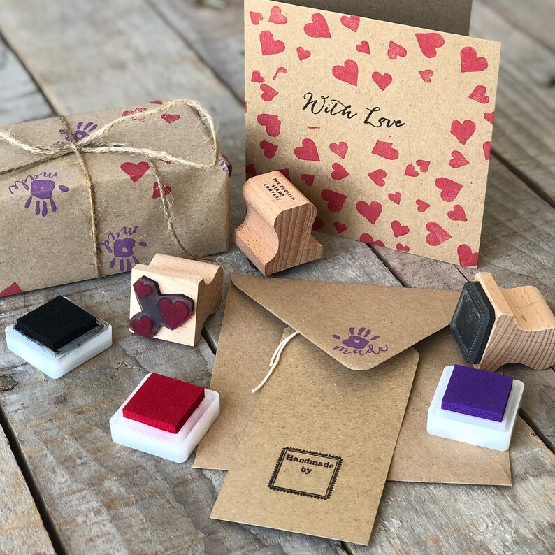 以下が含まれることがあります： ハート、手形、そして「With Love」と「Handmade by」の文字を含む様々なデザインのスタンプセット。スタンプは、様々な色のインクパッドと共に、茶色の紙の表面に配置されています。