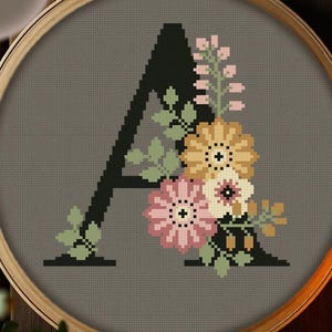 May include: A cross stitch pattern in a wooden embroidery hoop featuring a large black letter A decorated with pink, yellow, and white flowers and green leaves on a gray fabric background. The image includes the text PDF PATTERN in the corner.