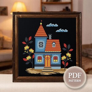 Pode incluir: Um padrão de ponto cruz numa moldura de madeira escura representando uma casa azul com telhado de telhas vermelhas, uma torre central e janelas iluminadas a amarelo. A casa é ladeada por árvores estilizadas com folhas vermelhas e amarelas.