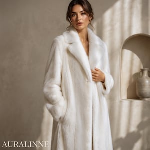 May include: A long, white faux fur coat with a classic collar and pockets. The coat is worn by a person. The brand name "AURALINNE" is visible at the bottom of the image. The coat appears to be made of a soft, plush material.