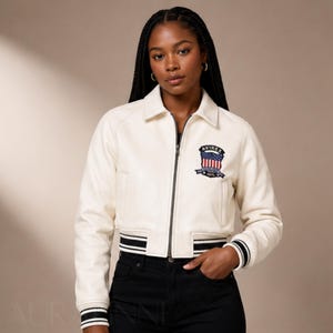 May include: Cream-colored leather jacket with a zip-up front, collar, and black and white striped cuffs and hem. Features a patch on the chest with the text "AVIREX" and an American flag design. The jacket is paired with black pants.