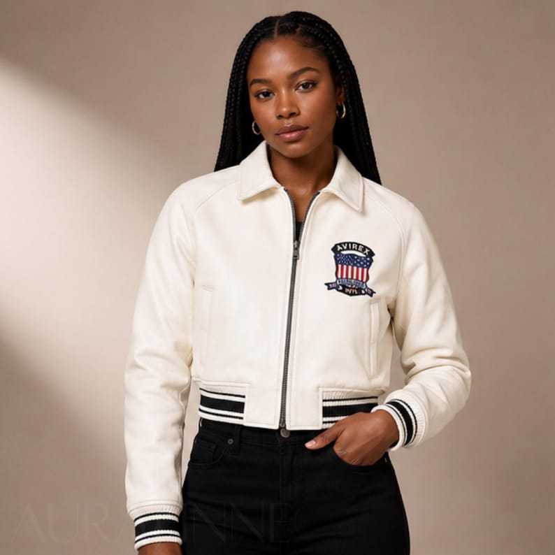 May include: Cream-colored leather jacket with a zip-up front, collar, and black and white striped cuffs and hem. Features a patch on the chest with the text "AVIREX" and an American flag design. The jacket is paired with black pants.