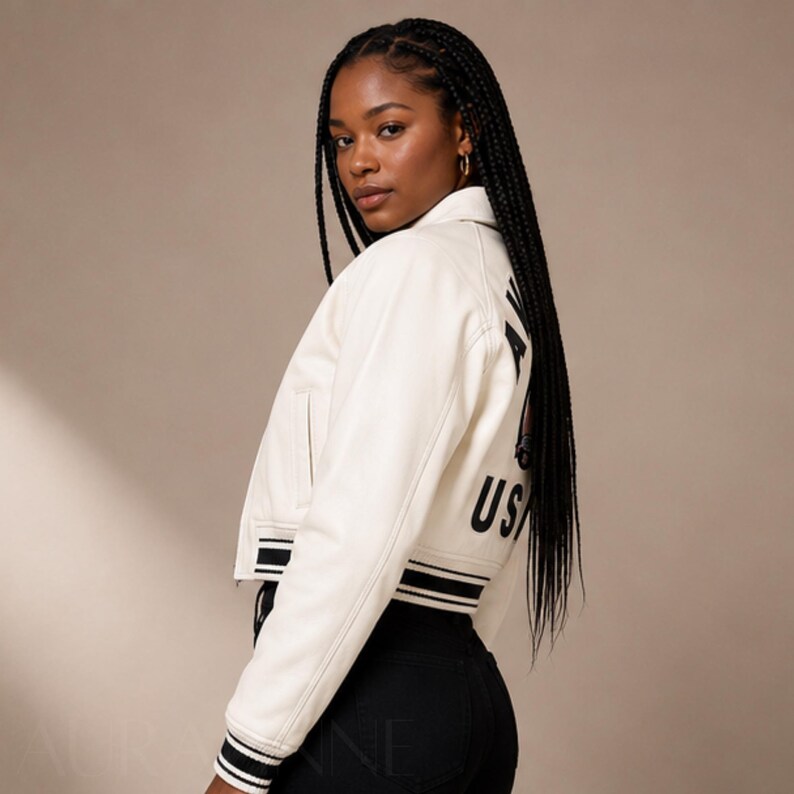 May include: Cream-colored cropped jacket with black and white striped trim. The jacket features the text "USA" in black on the back. The model is wearing black pants and has long black braids.