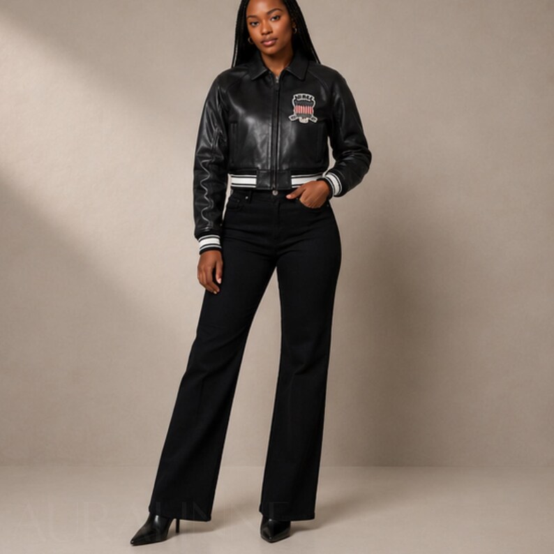 May include: A black leather jacket with a white and black striped trim on the cuffs and waist. The jacket has a patch on the chest. The model is wearing black flared jeans and black pointed-toe heels.