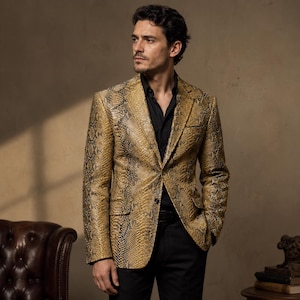 May include: A blazer featuring a tan and black snakeskin pattern with a notched lapel, two front flap pockets, and a single button closure. The garment is worn over a solid black button-down shirt and black trousers, creating a structured look.