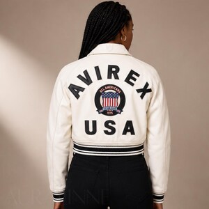 May include: Cream-colored cropped jacket with the word "AVIREX" in black block letters across the back. Below the word "AVIREX" is a circular patch with the American flag and the word "USA". The jacket has black and white striped trim.