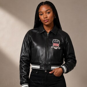 May include: Black leather bomber jacket with a white and black striped hem and cuffs. The jacket features a patch with the word "AVIREX" and a patriotic design. The jacket has a zipper closure and a collar.