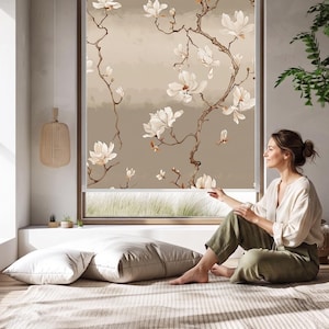 Pode incluir: Uma persiana de janela com um design floral de flores de magnólia brancas e galhos marrons em um fundo neutro. A persiana está instalada em uma sala com luz natural.