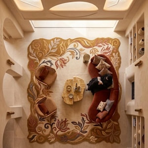 May include: An overhead view of a living room with a large, ornate rug featuring floral and leaf patterns in shades of gold, brown, and muted colors. A curved red sofa, two wicker chairs, and a unique wooden coffee table complete the space.