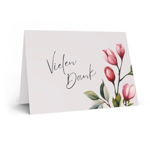 Könnte beinhalten: Eine gefaltete weiße Karte mit dem Schriftzug "Vielen Dank" in schwarzer Schrift. Die rechte Seite zeigt Aquarellillustrationen von rosa Blüten und grünen Blättern, die ein florales Design bilden.