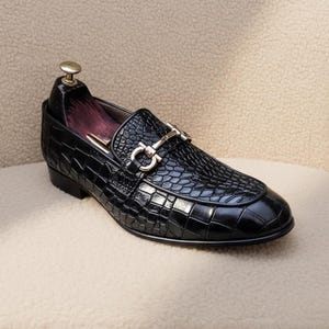 Può includere: Mocassino in pelle nera con texture a pelle di coccodrillo. La scarpa presenta una fibbia in metallo color argento e una punta arrotondata. La scarpa è su una superficie beige.