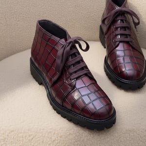 Könnte beinhalten: Burgunderfarbene Stiefeletten mit einem strukturierten, gitterartigen Muster. Die Stiefel haben dunkelbraune Schnürsenkel und dicke schwarze Sohlen. Die Schuhe werden auf einer cremefarbenen Oberfläche präsentiert.