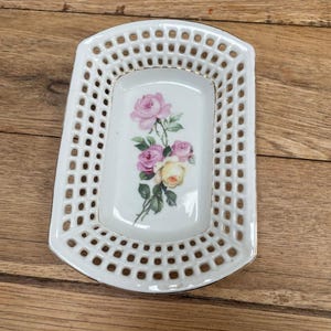May include: White ceramic dish with a rectangular shape and a pierced design. The center features a floral design with pink and yellow roses. The dish has a gold trim and is likely for decorative purposes.