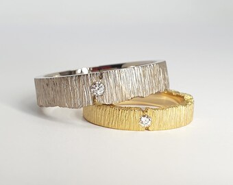 May include: Two textured metal rings, one silver and one gold, each with a small, clear gemstone. The rings have a bark-like texture and are set against a white background. The gold ring is stacked beneath the silver ring.