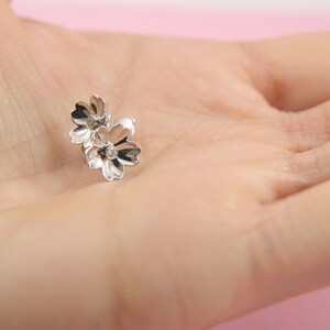May include: Silver flower stud earrings with a delicate design. The earrings feature two small flowers with a single rhinestone in the center of each flower.