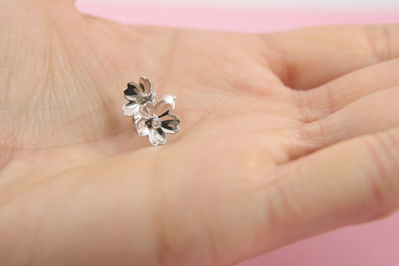 May include: Silver flower stud earrings with a delicate design. The earrings feature two small flowers with a single rhinestone in the center of each flower.