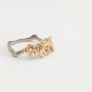 May include: A gold ring with a delicate floral design. The ring features three small flowers with white gemstones in the center. The band of the ring is made of a silver metal.