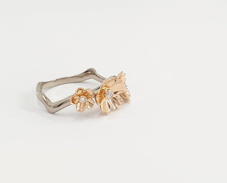 May include: A gold ring with a delicate floral design. The ring features three small flowers with white gemstones in the center. The band of the ring is made of a silver metal.
