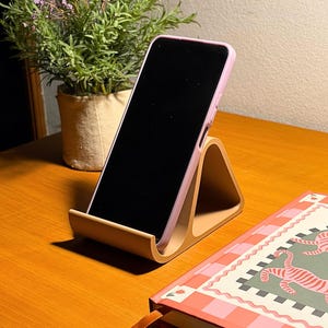 以下が含まれることがあります： ピンク色のケースに入った携帯電話を置く木製の電話スタンド。スタンドは、ピンクと赤のチェック柄のノートと植木鉢に入った植物の隣の木製の机の上にあります。
