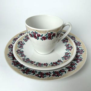 Puede incluir: Juego de té blanco con taza, platillo y plato. El juego presenta un estampado floral de rosas burdeos y verdes, con un borde dorado. La taza está sobre el platillo, que está encima del plato.