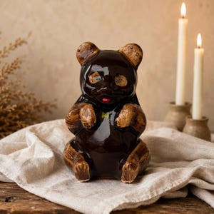 Może przedstawiać: Błyszcząca, ciemnobrązowa ceramiczna figurka niedźwiedzia z jasnobrązowymi akcentami na uszach, łapach i stopach. Niedźwiedź ma małe zielone oczy, czerwone usta i mały zielony detal na piersi, przedstawiony w pozycji siedzącej z uniesionymi ramionami.