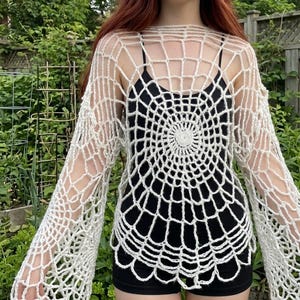 May include: A white long-sleeve crochet top featuring an intricate, circular spiderweb pattern that radiates from the center. The garment is constructed with an open-knit design, creating a net-like texture across the sleeves and torso.