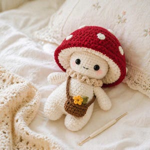 May include: A handmade crochet mushroom doll with a red cap dotted with white spots. The doll has a cream-colored body, a smiling face, and a small brown purse with a yellow flower. A crochet hook rests nearby.